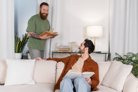 Positive gay couple holding books while resting in modern living room 