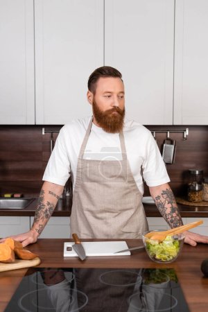 Tattooed man in apron standing near fresh salad in kitchen 