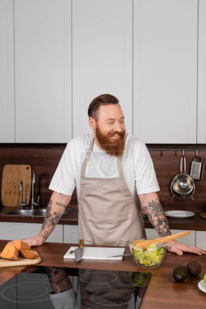 Alegre hombre tatuado en delantal de pie cerca de la comida y ensalada fresca en la cocina 