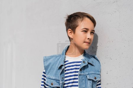 well dressed preteen boy in denim vest and striped long sleeve shirt standing near mall with grey wall