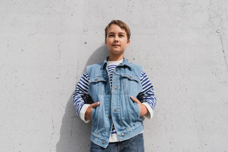 stylish preteen boy in denim vest and striped long sleeve shirt posing with hands in pockets near mall with grey wall