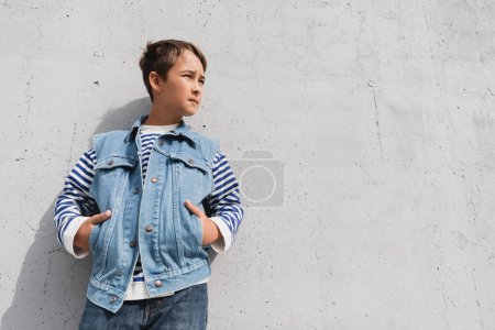 low angle view of stylish boy in denim vest and striped long sleeve shirt posing with hands in pockets near mall with grey wall