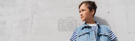 low angle view of stylish boy in denim vest and striped long sleeve shirt looking away near mall with grey wall, banner 