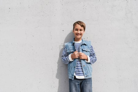 Foto de Niño feliz en chaleco de mezclilla y camisa de manga larga a rayas de pie cerca del centro comercial con pared gris - Imagen libre de derechos