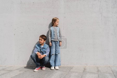 in voller Länge stylische Kinder in Jeans-Outfits mit gestreiften Langarmhemden, die in der Nähe der grauen Wand in einem Einkaufszentrum posieren 