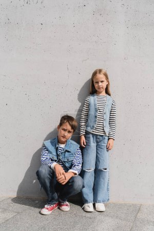 full length of stylish preteen kids in denim outfits posing near grey wall in mall 