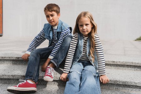 Photo pour Well dressed children in denim vests with long sleeve shirts sitting on stairs near mall - image libre de droit