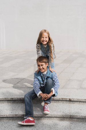 Photo pour Cheerful girl in stylish clothes leaning on shoulders of happy preteen boy sitting on stairs - image libre de droit