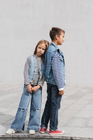 full length of well dressed girl in denim outfit leaning on back of boy while standing outdoors 