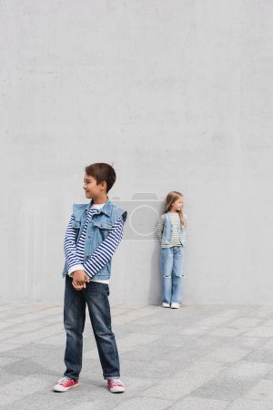 volle Länge des fröhlichen Jungen in stilvollem Jeans-Outfit, der neben Mädchen auf verschwommenem Hintergrund steht 