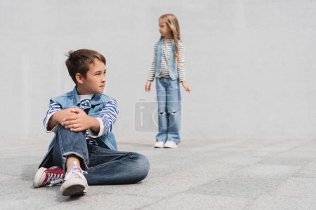 full length of well dressed boy in denim outfit sitting near girl on blurred background 