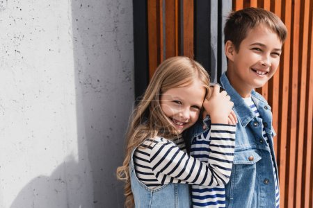 Porträt fröhlicher Kinder in gestreiften langärmeligen Hemden und Jeanswesten, die in der Nähe eines Gebäudes in die Kamera schauen 