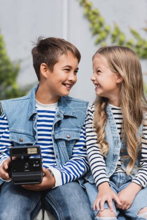 Téléchargez les photos : Heureux garçon préadolescent dans des vêtements élégants tenant caméra vintage près de fille bien habillée - en image libre de droit