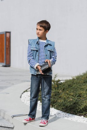 full length of well dressed preteen boy in denim clothes holding vintage camera outside 
