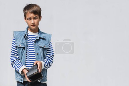 well dressed preteen boy in denim clothes holding vintage camera on grey background  