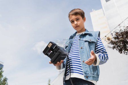 low angle view of well dressed preteen boy in denim clothes holding vintage camera outside 