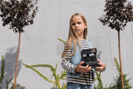 niña en chaleco de mezclilla azul y camisa de manga larga a rayas que sostiene la cámara vintage cerca del edificio del centro comercial 