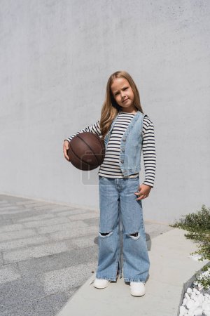 full length of preteen girl in denim vest and blue jeans holding basketball near mall building 