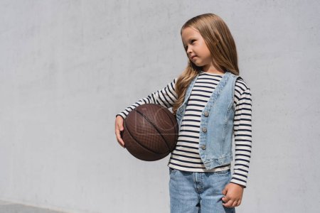 preteen girl in denim vest and blue jeans holding basketball near mall building 