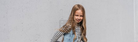 Photo for Cheerful girl in denim vest striped long sleeve shirt standing near grey wall, banner - Royalty Free Image