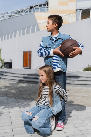 girl in stylish clothes hugging legs of boy with basketball standing near mall 