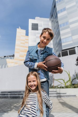 Photo pour Cheerful boy in denim vest holding basketball near smiling girl while standing near mall - image libre de droit