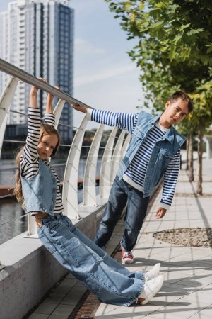 Photo pour Full length of stylish kids in denim vests and jeans posing near metallic fence on riverside - image libre de droit