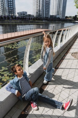 Blick aus der Vogelperspektive auf gut gekleidete Kinder in Jeanswesten und Jeans, die in der Nähe eines Metallzauns am Ufer des Flusses posieren 