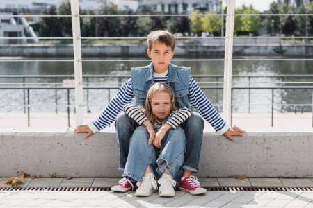 Stylische Kinder in Jeanswesten und Jeans sitzen in der Nähe von Metallzaun am Flussufer