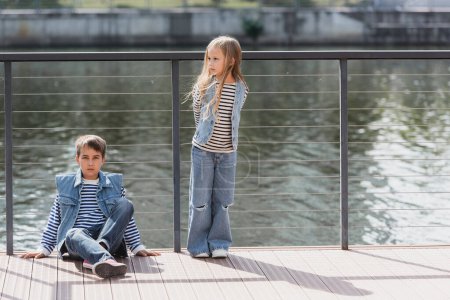 In voller Länge posieren gut gekleidete Kinder in Jeanswesten und Jeans neben Zaun am Flussufer