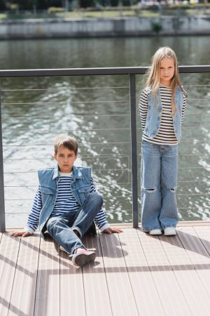 In voller Länge posieren gut gekleidete Kinder in Jeanswesten und Jeans neben Zaun am Flussufer 