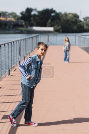 full length of preteen boy in denim outfit posing near girl standing on river embankment on blurred background 