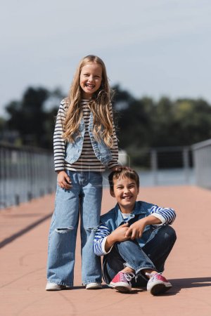 full length of smiling girl in denim outfit standing near happy preteen boy on river embankment 