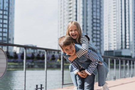 happy preteen boy in denim outfit piggybacking girl on river embankment 