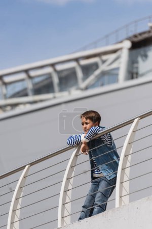 low angle view of preteen boy in denim vest and jeans leaning on metallic fence on embankment 