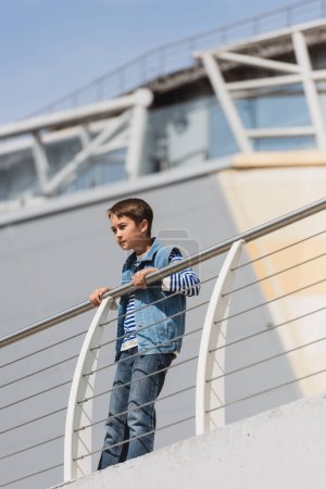 vista de ángulo bajo de niño con estilo en chaleco de mezclilla y jeans de pie cerca de valla metálica en el terraplén 