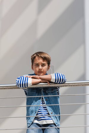 stylish boy in denim vest and long sleeve shirt standing near metallic fence on embankment 