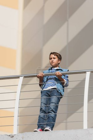 Foto de Vista de ángulo bajo de niño con estilo en chaleco de mezclilla y jeans de pie cerca de valla metálica - Imagen libre de derechos