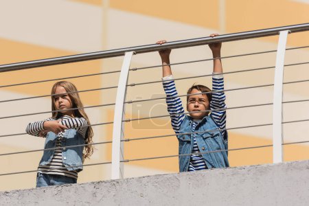 low angle view of stylish children in denim vests and striped long sleeve shirts standing near metallic fence 