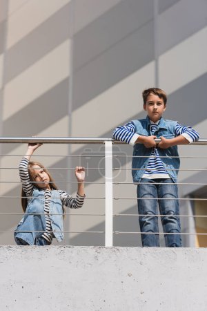 low angle view of children in stylish denim vests and striped long sleeve shirts standing near metallic fence 