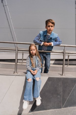 stylish kids in casual denim attire posing near metallic handrails next to building 