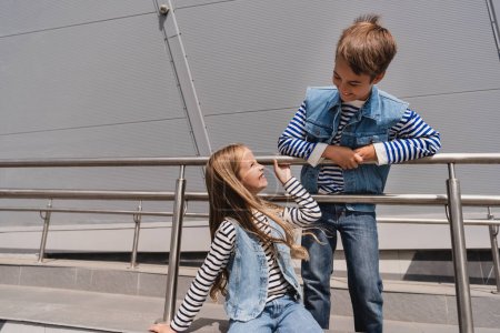 glückliche und gut gekleidete Kinder in lässiger Jeans-Kleidung posieren in der Nähe von Metallgeländern neben dem Gebäude 