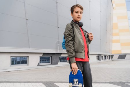 Téléchargez les photos : Preteen boy in bomber jacket holding penny board while standing near mall - en image libre de droit