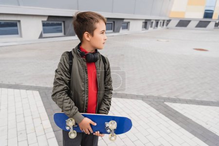 Foto de Preteen boy in bomber jacket and wireless headphones holding penny board while standing near mall building - Imagen libre de derechos