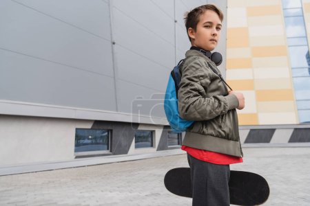 preteen boy in wireless headphones standing with backpack and holding penny board near mall building 
