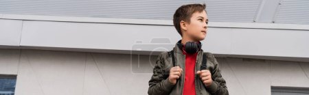 preteen boy in bomber jacket and wireless headphones holding backpack while standing near mall, banner 