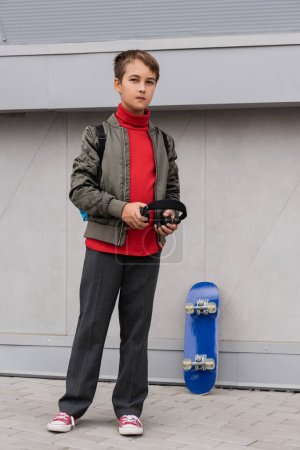 preteen boy in bomber jacket holding wireless headphones while standing near penny board next to mall 