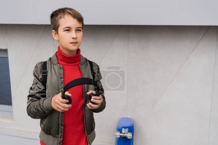 Foto de Preteen boy in bomber jacket holding wireless headphones while standing near penny board next to mall building - Imagen libre de derechos