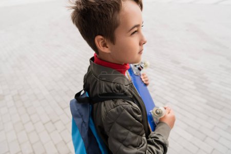 preteen boy in stylish bomber jacket standing with backpack while holding penny board 