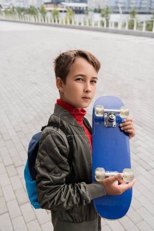 preteen boy in trendy bomber jacket standing with backpack while holding penny board 
